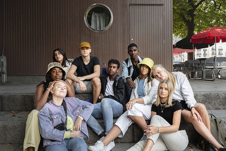 En grupp av ungdomar med olika etnisk bakgrund som sitter i en trappa utomhus. Brun trävägg bakom dem.