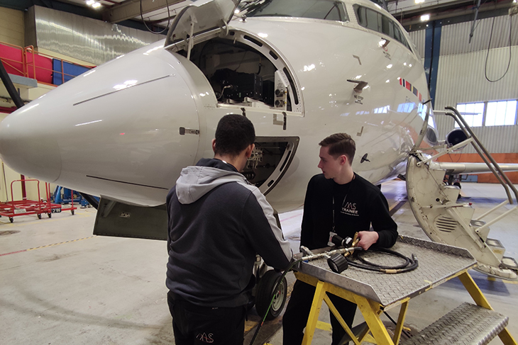 Två killar står vid ett flygplan i en hangar med mekaniska instrument. Flygplanets nos är öppen och ska repareras.