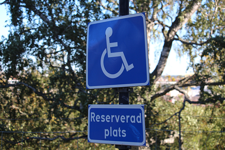 En blå trafikskylt med en handikappsymbol och texten Reserverad plats under. I bakgrunden gröna träd. 