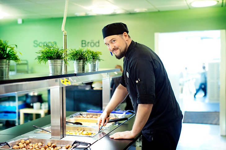En man i svarta kockkläder står vid en serveringsdisk i en skolrestaurang