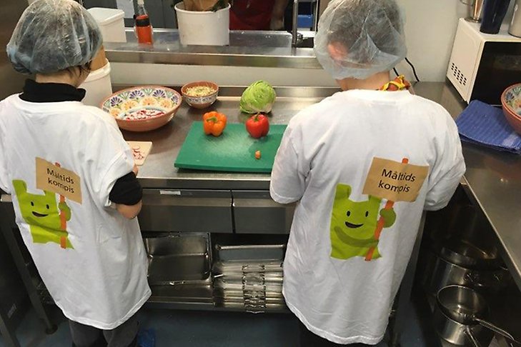 Två barn står och skär grönsaker. Man ser ryggarna. De har likadana t-shirtar på sig där det står måltidskompis.