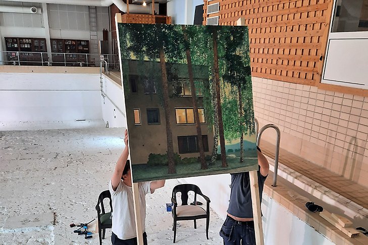 Två personer lyfter en tavla med en målning av ett flerbostadshus och mycket grönska.