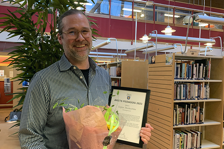 En man i rutig skjorta står med diplom och blommor i handen i ett bibliotek med bokhyllor bakom sig. 