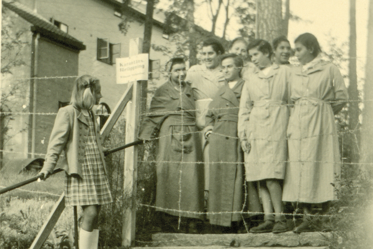 Svartvitt foto på ett flertal unga kvinnor som står på ena sidan av ett stängsel. En annan ung kvinna står på andra sidan. De ler mot varandra.