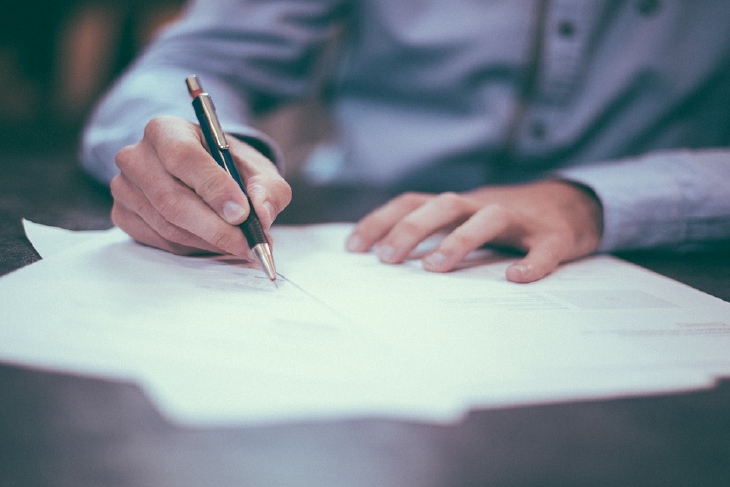 En man i blå skjorta håller i en bläckpenna och
skriver på ett par papper som ligger på bordet framför honom