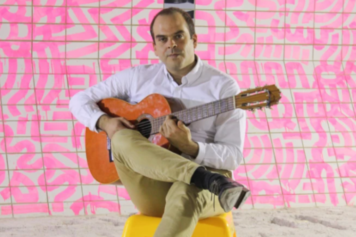En man med gitarr i knät sitter på en stor och tittar rakt fram.