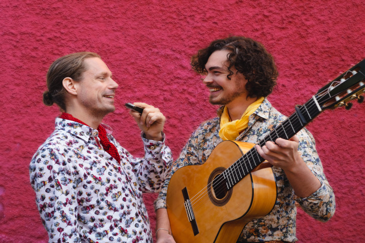 Två män med gitarr och munspel tittar på varandra och skrattar.