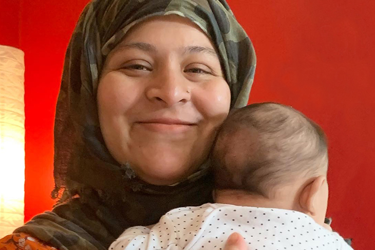 En kvinna med ett spädbarn i famnen ler och tittar in i kameran. Hon har en mörkgrön hijab på huvudet.