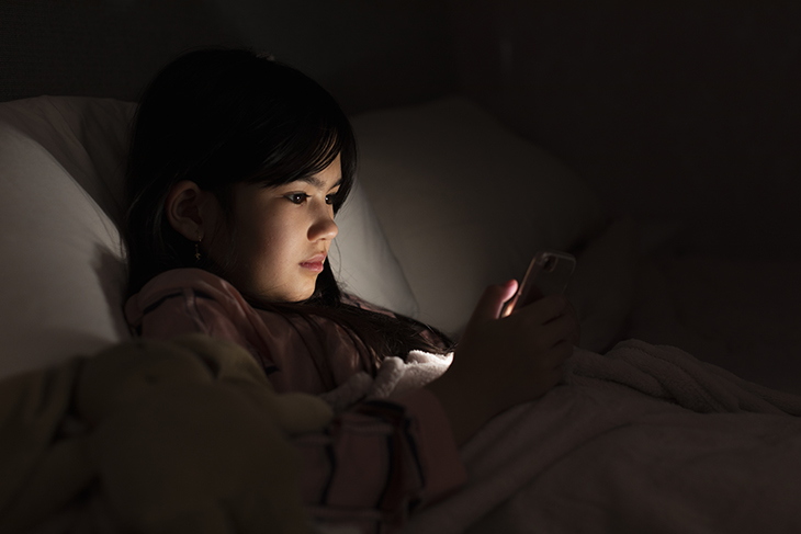 Mörkt sovrum. En tonårstjejs ansikte lyses upp av ljus från mobilen. 