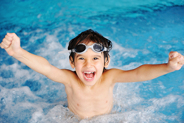 En pojke badar i en pool med simglasögon på huvudet och ler stort med munnen öppen och armarna sträckta i luften.