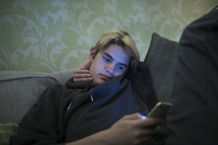 Tonårig kille ligger i en soffa och tittar på sin mobiltelefon. Han ser allvarlig och lugn ut. 