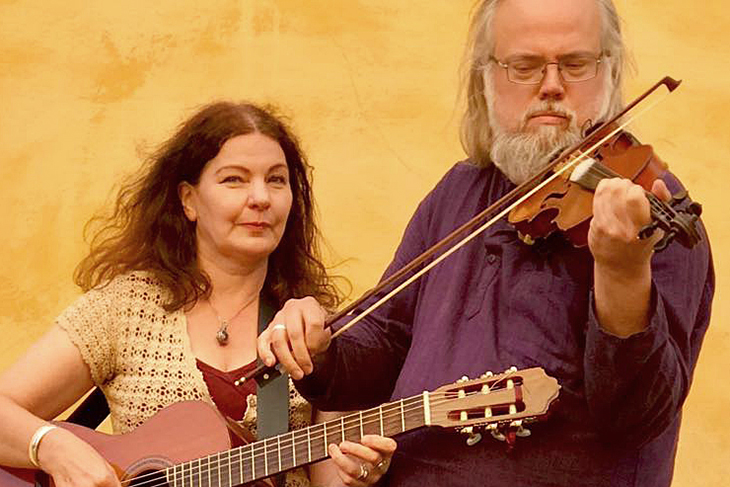En kvinna spelar gitarr och en man spelar fiol mot en gul vägg.