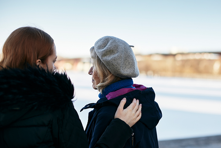 En kvinna håller sin hand om ryggen på en äldre dam. De står utomhus och det är vinter. 