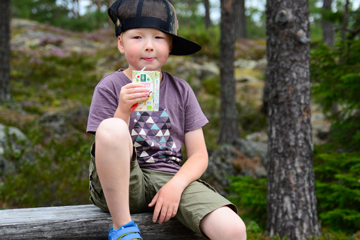 Pojke i t-shirt och keps på utflykt i skogen dricker saft ur tetra. 