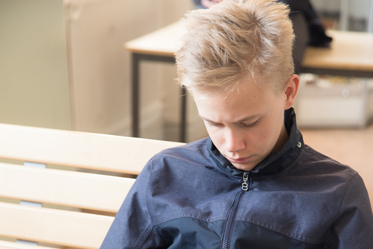 Blond kille sitter på en bänk i sin skola och tittar i sin mobiltelefon. Ser ledsen ut. 