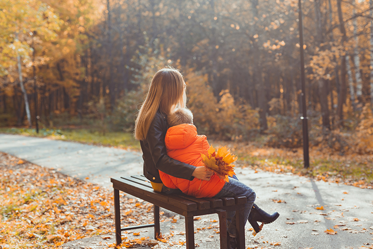 Kvinna med långt hår och svart jacka sitter på en parkbänk och håller om en pojke i tioårsåldern i orange jacka. Kvinnan håller en bukett med lönnlöv i höstfärger. 