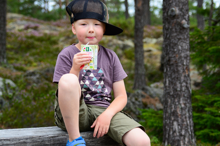 Pojke i svart keps, t-shirt och shorts sitter vid en rastplats i skogen och dricker äppeljuice ur en tetra. 