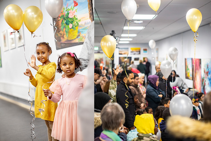 Två unga flickor klädda i gul och rosa klänningar står framför konstverk och vinkar med ballonger i ena handen. Folkmassa står framför samma konstverk och ballonger hänger i taket.