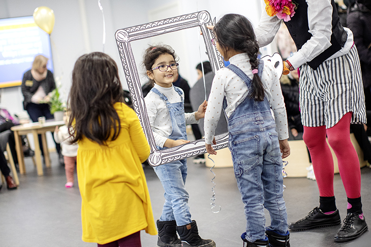 Två flickor iklädda liknande kläder ser på varandra genom en tecknad spegelram som en clownklädd person håller mellan dem.
