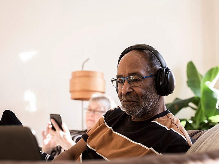 En äldre man med hörlurar sitter i en soffa och tittar på en datorskärm. Mannen har kort skägg, ganska mörk hudton och randig tröja. Bakom honom i soffan sitter en kvinna och tittar på sin mobiltelefon. 