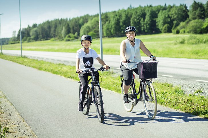 En vuxen och ett barn på cykel, cyklandes på cykelväg med ett fält i bakgrunden.