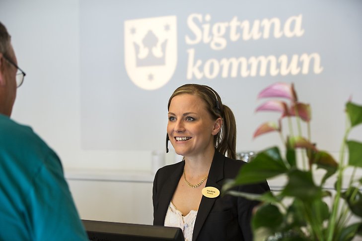 Receptionen på Sigtuna kommunhus. Leende receptionist och besökare med Sigtuna kommun logotype i bakgrunden. 
