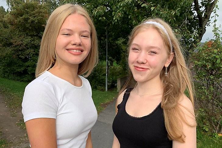 Två blonda flickor som går i högstadiet står bredvid varandra. Den ena har vit t-shirt, den andra ett svart linne. De ler in i kameran. Gröna träd i bakgrunden.