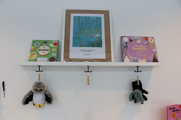 En hylla med två barnböcker. Mittemellan står en inramad bild med citat om barnböcker. På krokarna under hyllan hänger två handdockor i form av djur.