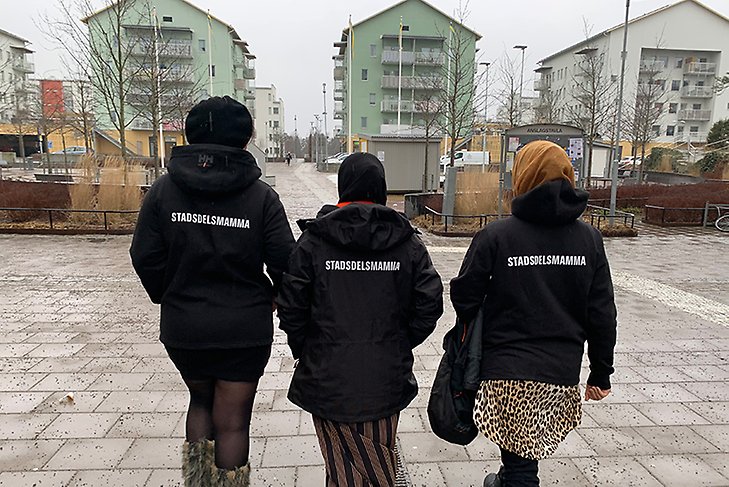 Tre kvinnor i svarta jackor med ordet Stadsdelsmamma i vit text på ryggen. De går i en stadsmiljö med höghus längst bort i bild. 