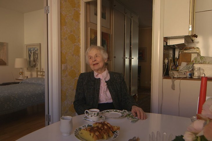 En äldre dam sitter och fikar i ett kök.