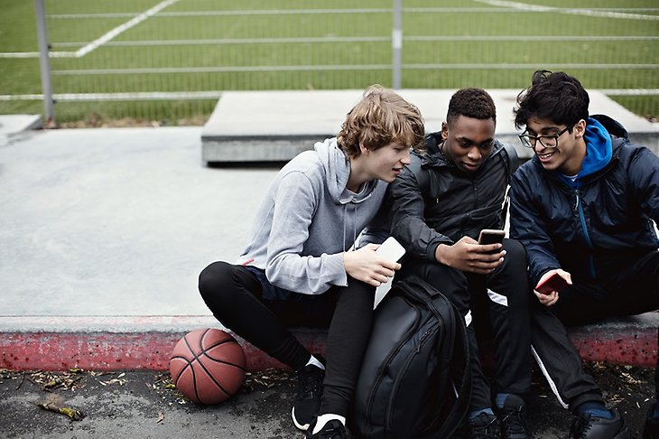 Tre unga killar kollar tillsammans på en mobiltelefon sittandes vid en idrottsplats bredvid en basketboll. 