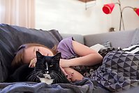 Flicka i lila tröja sover i en soffa. En svartvit katt ligger bredvid.