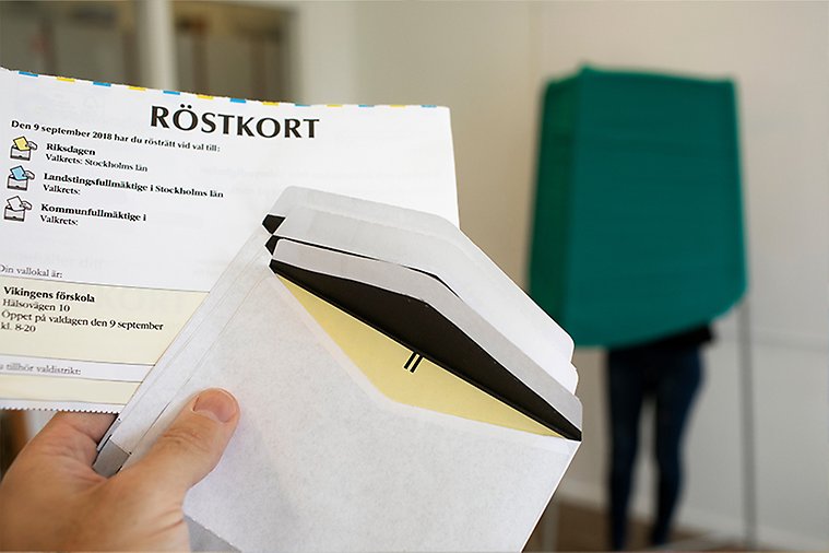 En hand håller upp ett röstkort och tre små kuvert framför ett röstningsbås. Foto: Plattform/Johnér