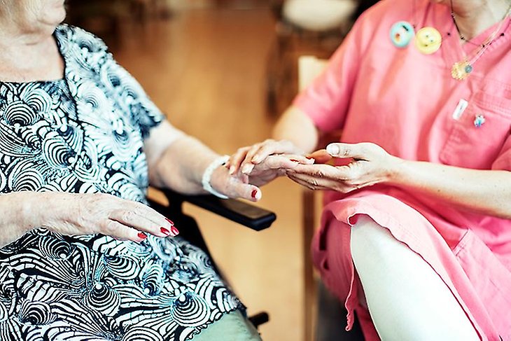 Äldre kvinna i rullstol sida vid sida med en undersköterska som håller hennes hand.