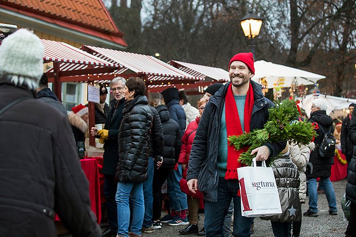 Människor som handlar i marknadsstånd. En man står vänd mot kameran och ler med en granrisklädd julbock i famnen. 