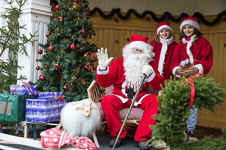En jultomte i röd dräkt och vitt skägg sitter i en fåtölj med gran, julklappar och granrisdekorationer brevid sig. Två barn i tomtenissedräkter står bakom. 