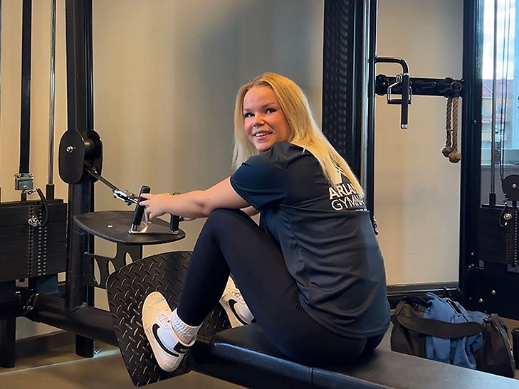 Ljushårig tjej i svart träningströja med Arlandagymnasiets logotyp på ryggen tränar i en maskin på gym. Hon vänder sig om och tittar in i kameran. 