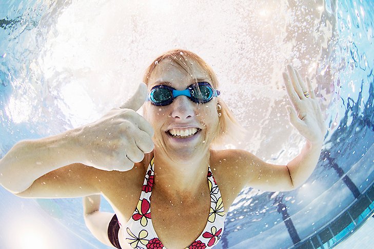 En kvinna iklädd badkläder och simglasögon gör tumme upp under vattenytan i en simbassäng.