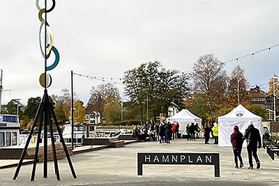 Högt konstverk bredvid skylt med text "Hamnplan" och människor som går mot partytält lite längre bort i höstkylan vid hamnen.