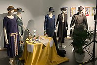 Bilder på skyltdockor i tidsenliga kläder. Ett bord med en gul duk och kaffekoppar står synligt i bilden.