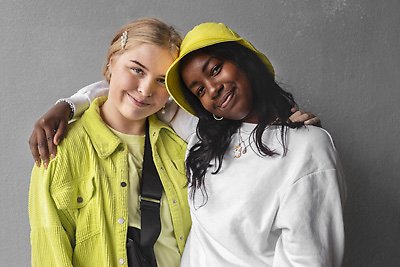 Två tonåringar iklädda gula och vita kläder håller om varandra och ler in i kameran framför en betongvägg.