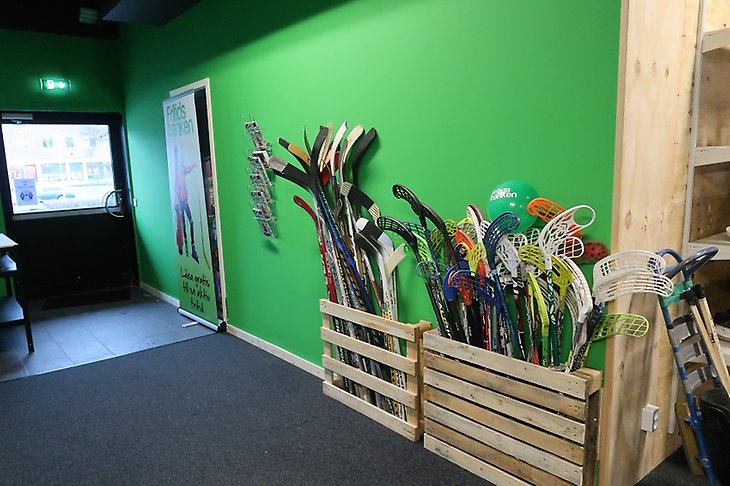 Entré till en lokal med grönmålade väggar och hockey- och innebandyklubbor längs väggen.