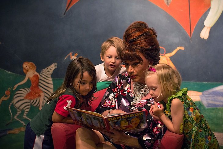 En kvinna med stor peruk läser högt ur en barnbok för tre barn runt om henne.