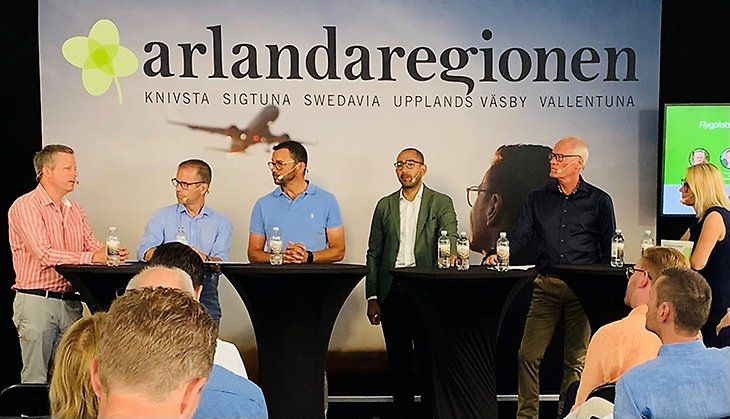 Sex personer står vid ståbord på en scen. Banderoll i bakgrunden där det står Arlandaregionen.